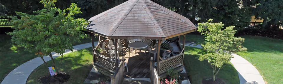 A wooden gazebo with a shingled roof situated in a green outdoor area with a curved concrete pathway and small trees surrounding it. Inside the gazebo, there are several chairs and a round table.