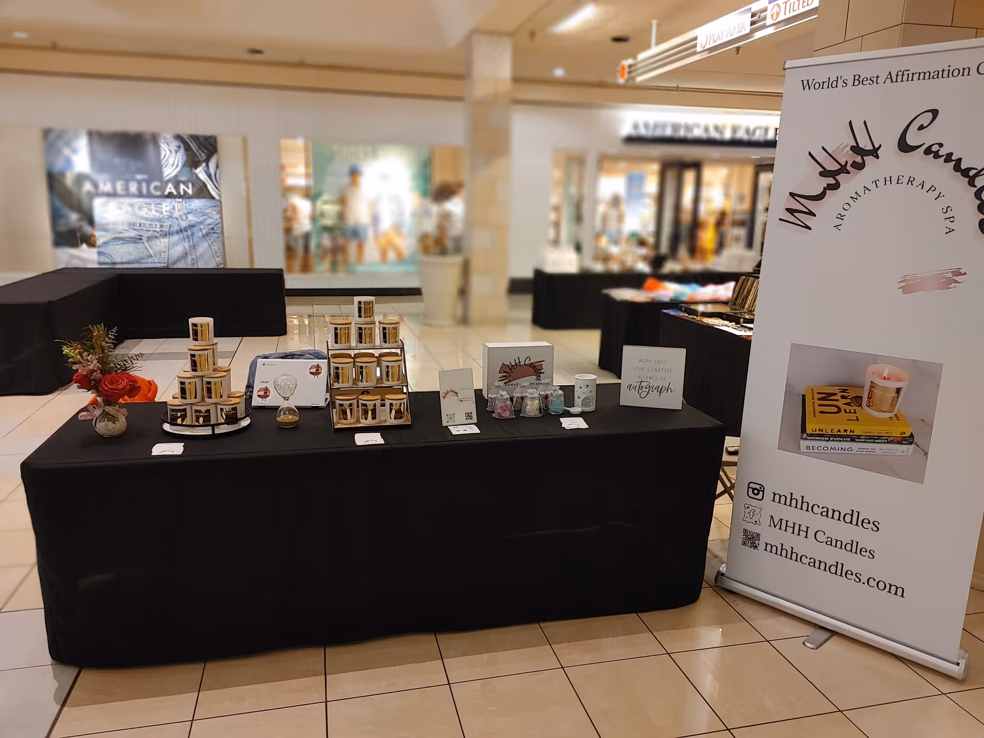 Indoor mall display featuring black-draped tables and a tall banner for MHH Candles.