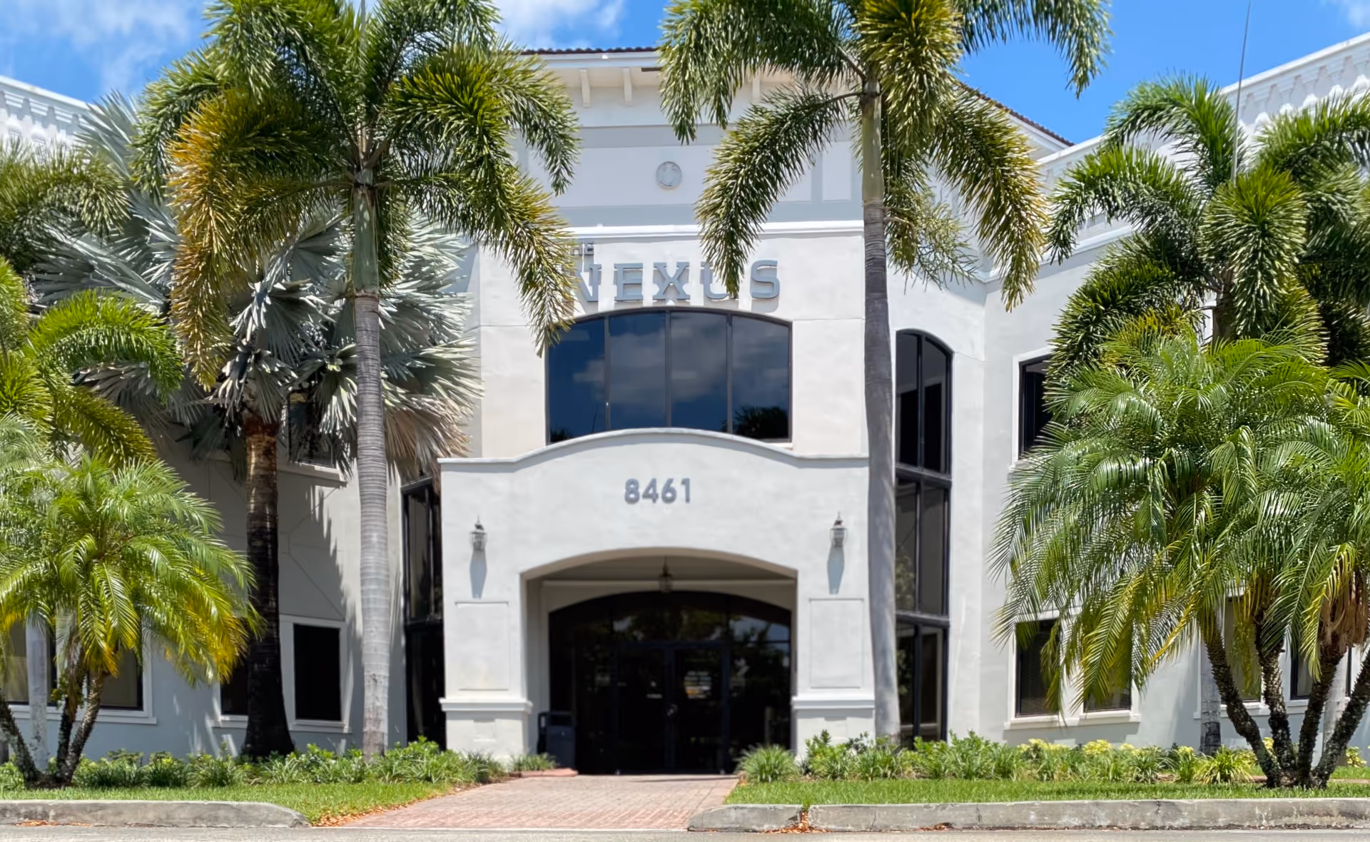 Front entrance of a light-colored building with palm trees and a curved archway.