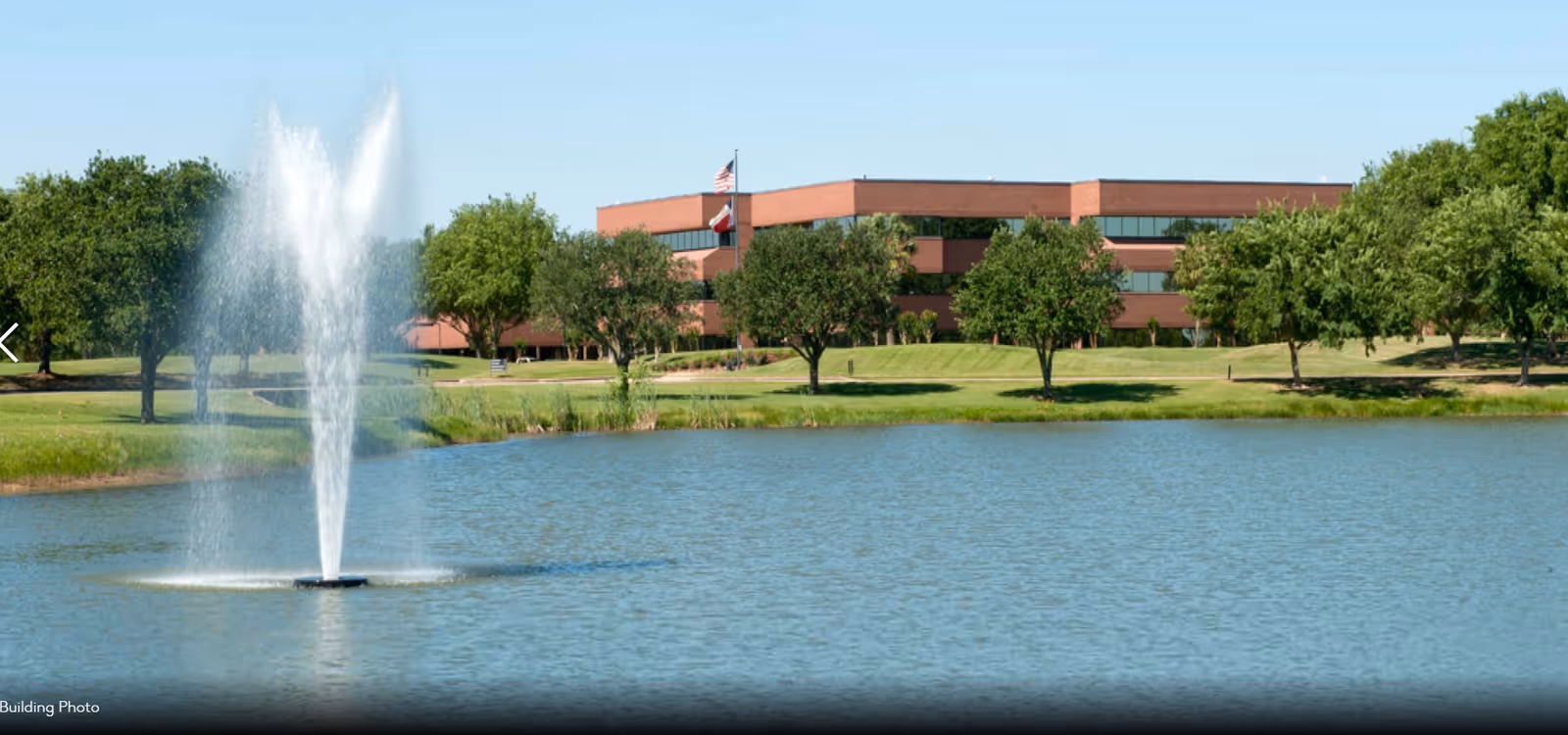 A brick campus building sits across a lake with a tall water fountain and green trees.