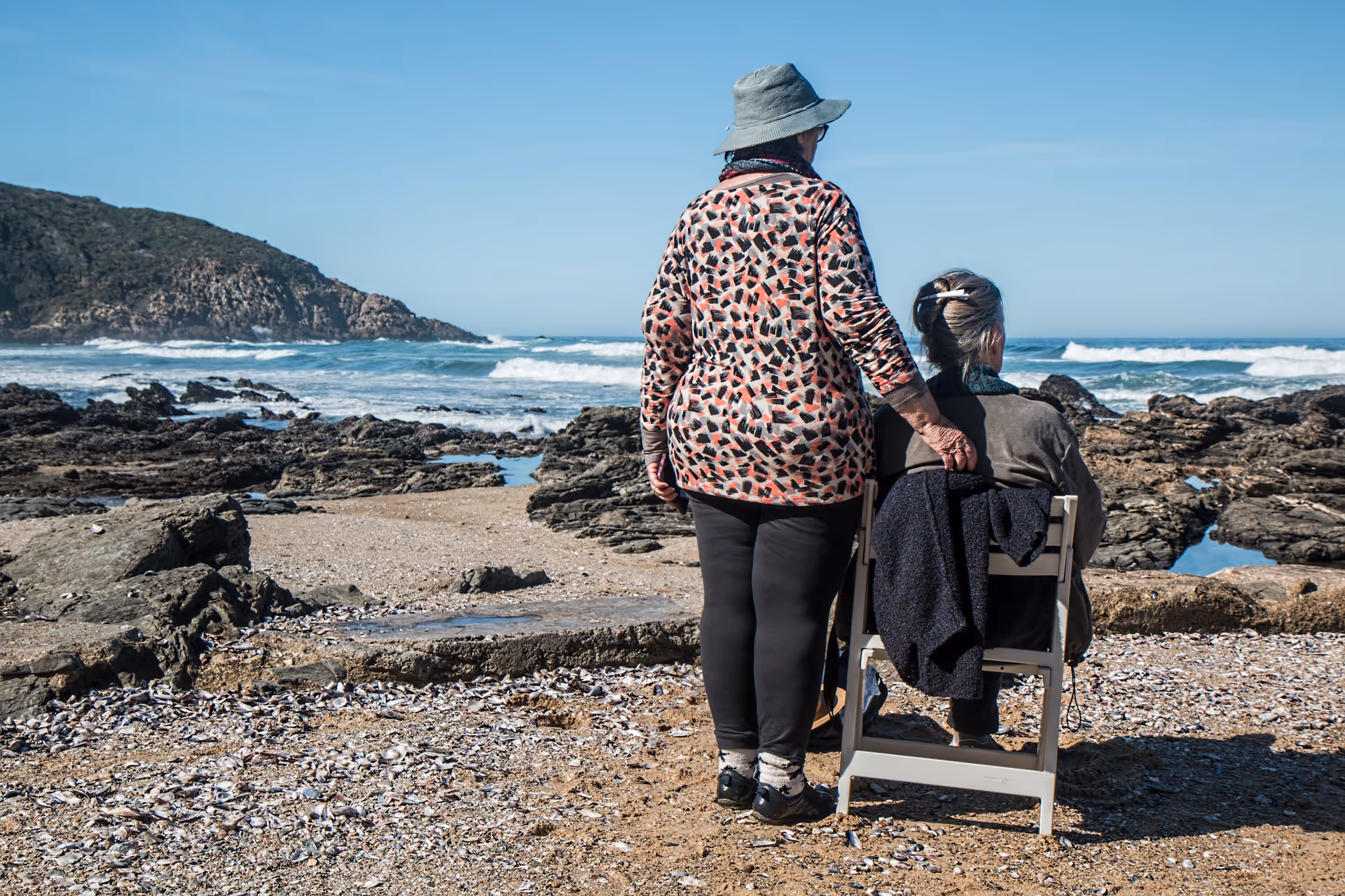 Two elderly individuals on a rocky beach, one standing beside a seated woman.