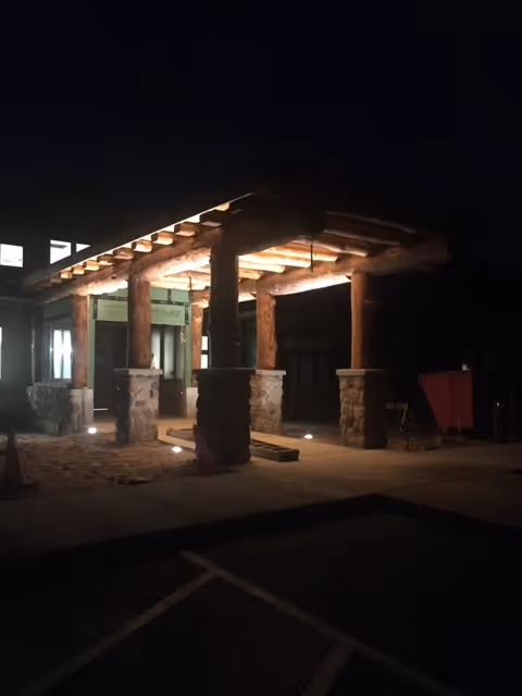Illuminated wooden porte-cochère with log beams and stone pillars at the front entrance of a building at night.