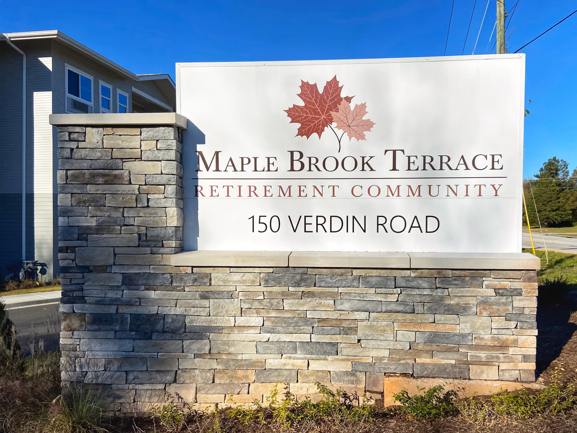 Stone and concrete sign for Maple Brook Terrace Retirement Community at 150 Verdin Road with a building and clear blue sky in the background.