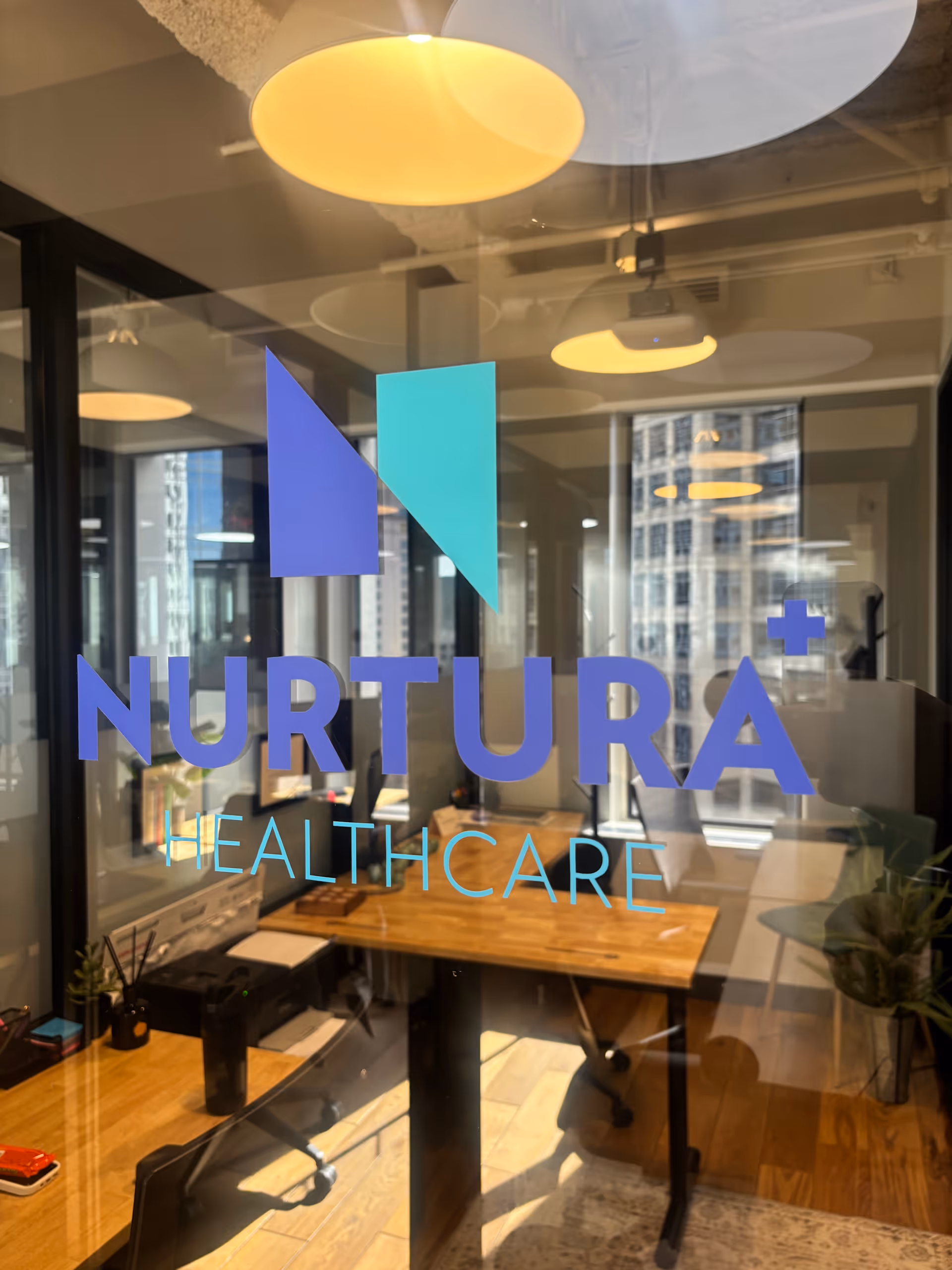 Glass door displaying Nurtura Healthcare logo over an office desk and city view.