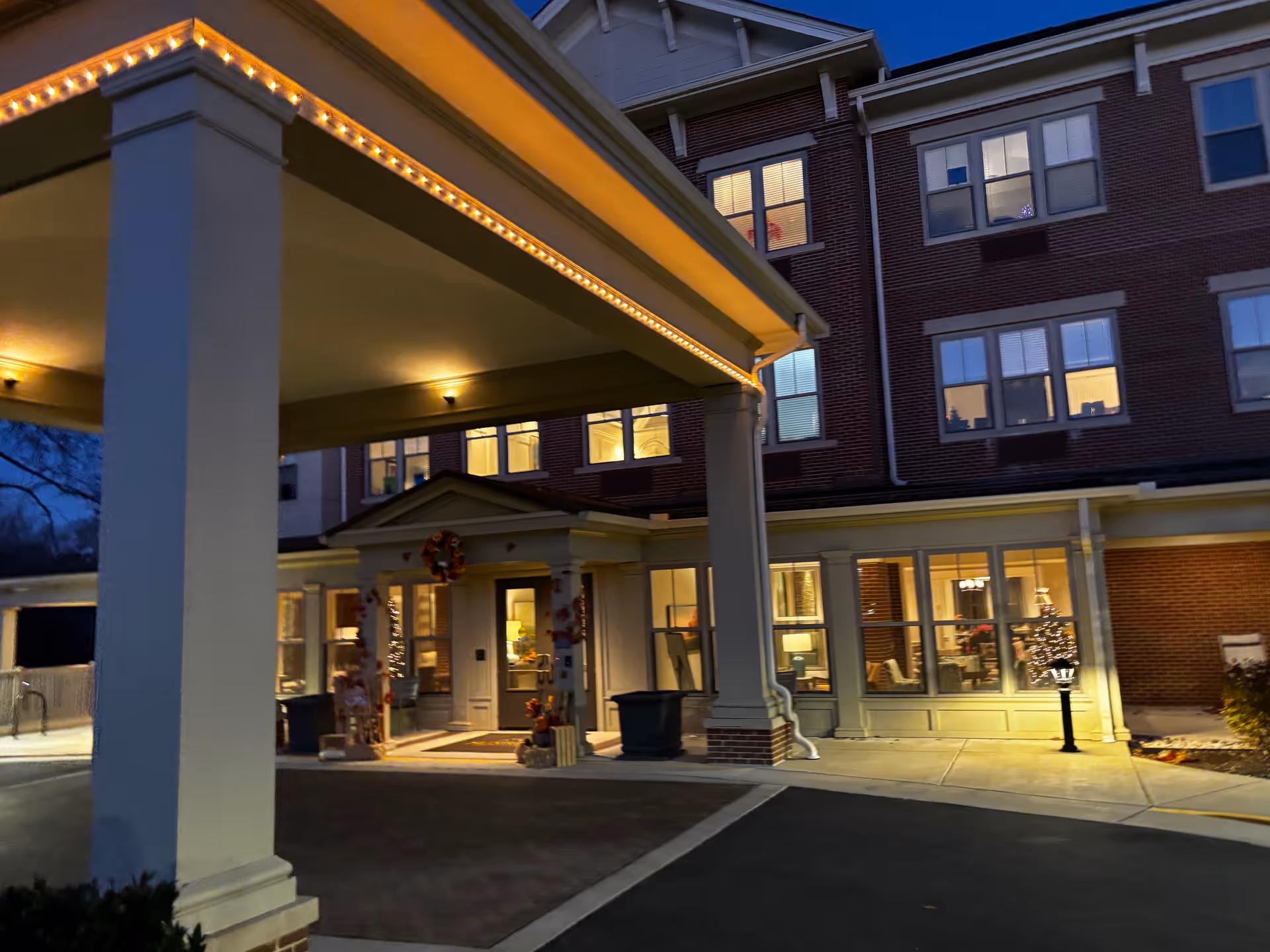 Front entrance and covered porte-cochere of the Sunrise of Fairfax senior living building lit at dusk with holiday decorations.