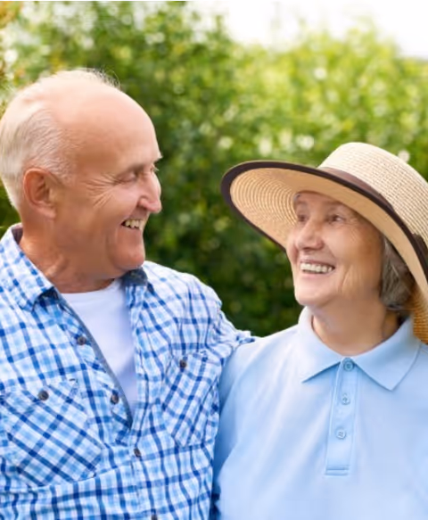 Two smiling seniors outdoors, wearing casual clothes, enjoying each other's company with greenery in background.