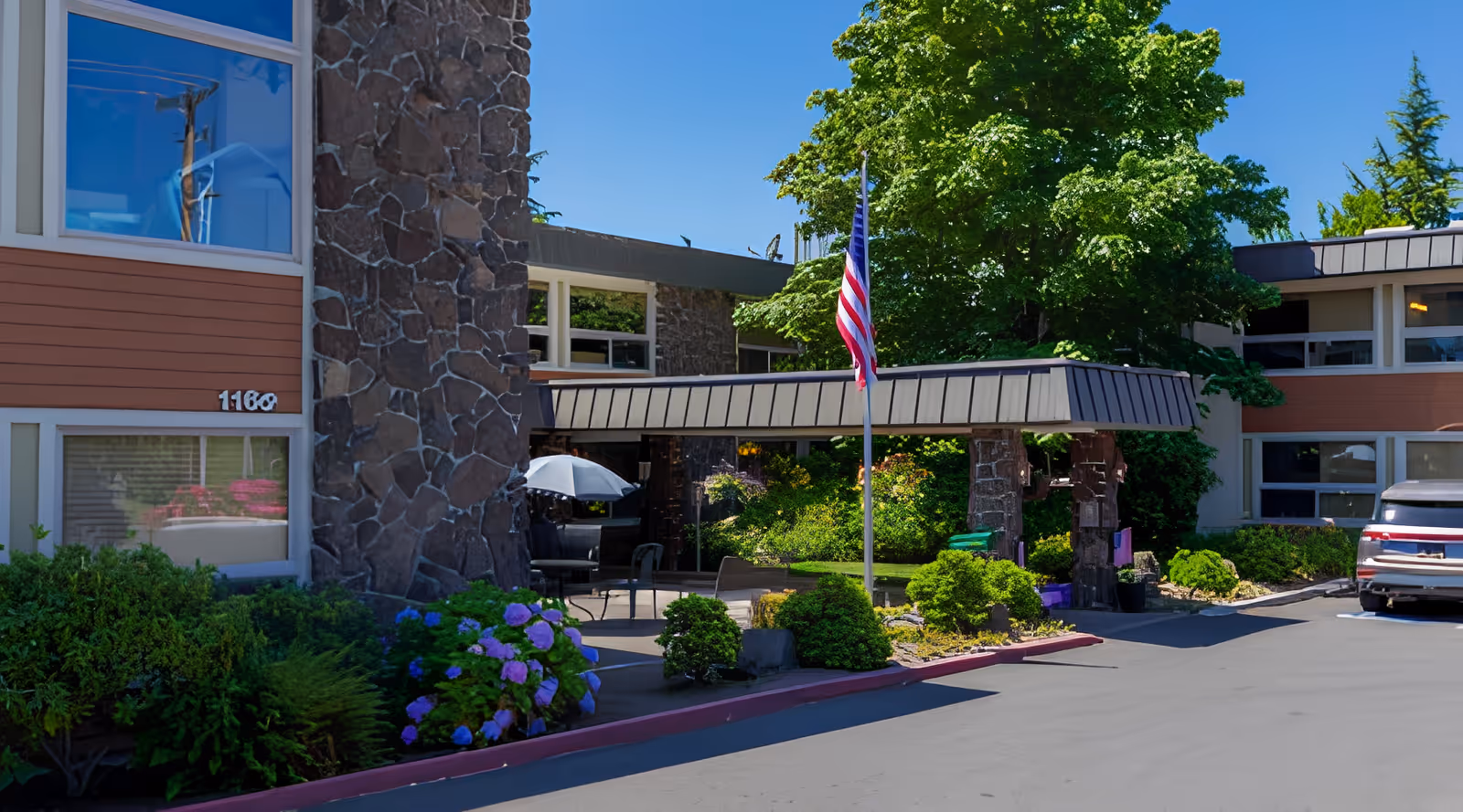 Front exterior of a senior living facility showing a stone-faced building with a covered entrance, American flag, landscaping, and a parked vehicle.