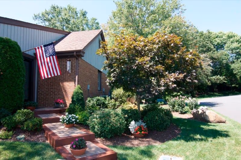 Front entrance of a brick building with an American flag, landscaped steps, potted flowers and a small tree.