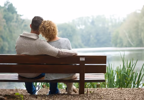 Two people sit on a wooden bench by a lake, surrounded by trees.