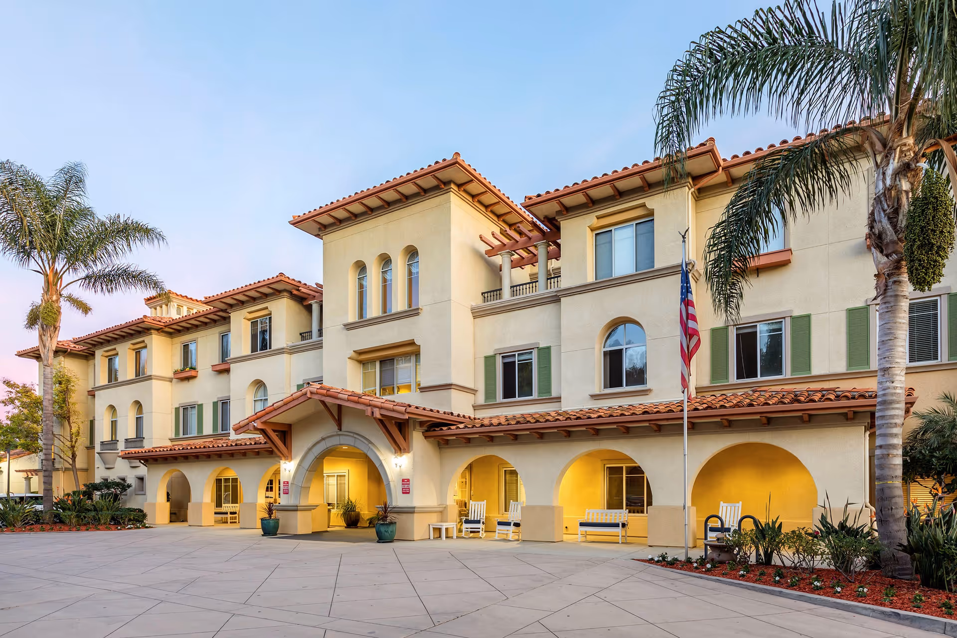Multi-story Mediterranean-style senior living building with arches, palm trees, and a flag at the entrance