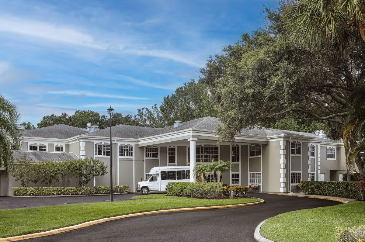 Entrance and front exterior of a senior living facility with a covered portico, shuttle van, and landscaped circular driveway.