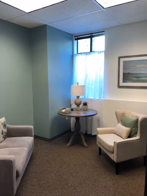 A small lounge with two chairs, a sofa, round table, and a lamp near a window.