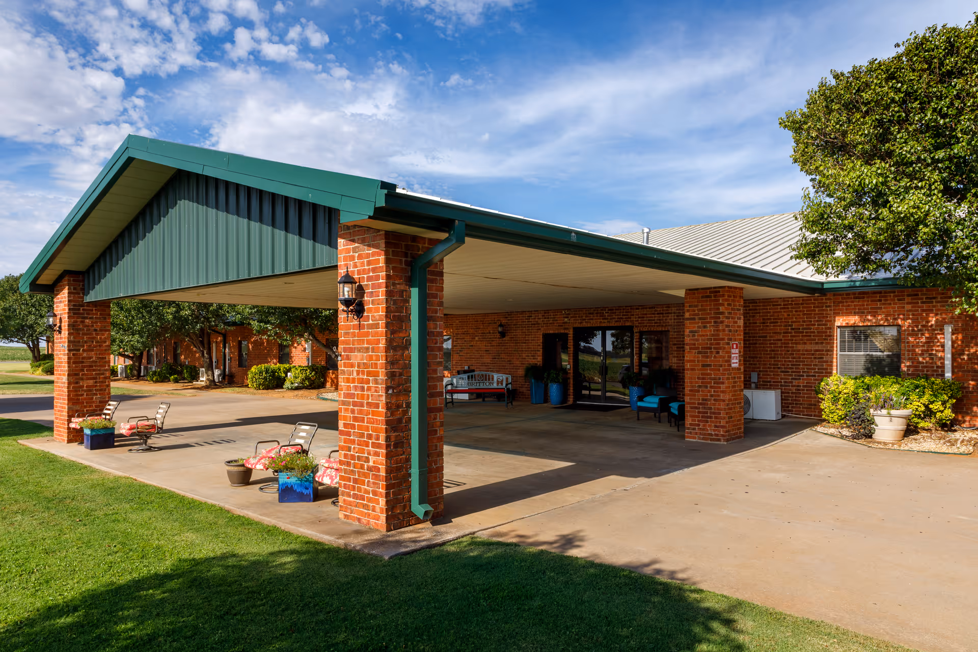 Covered entrance area of a brick building with green roof trim, outdoor seating with chairs and potted plants, and a concrete driveway surrounded by green grass and trees under a partly cloudy sky.