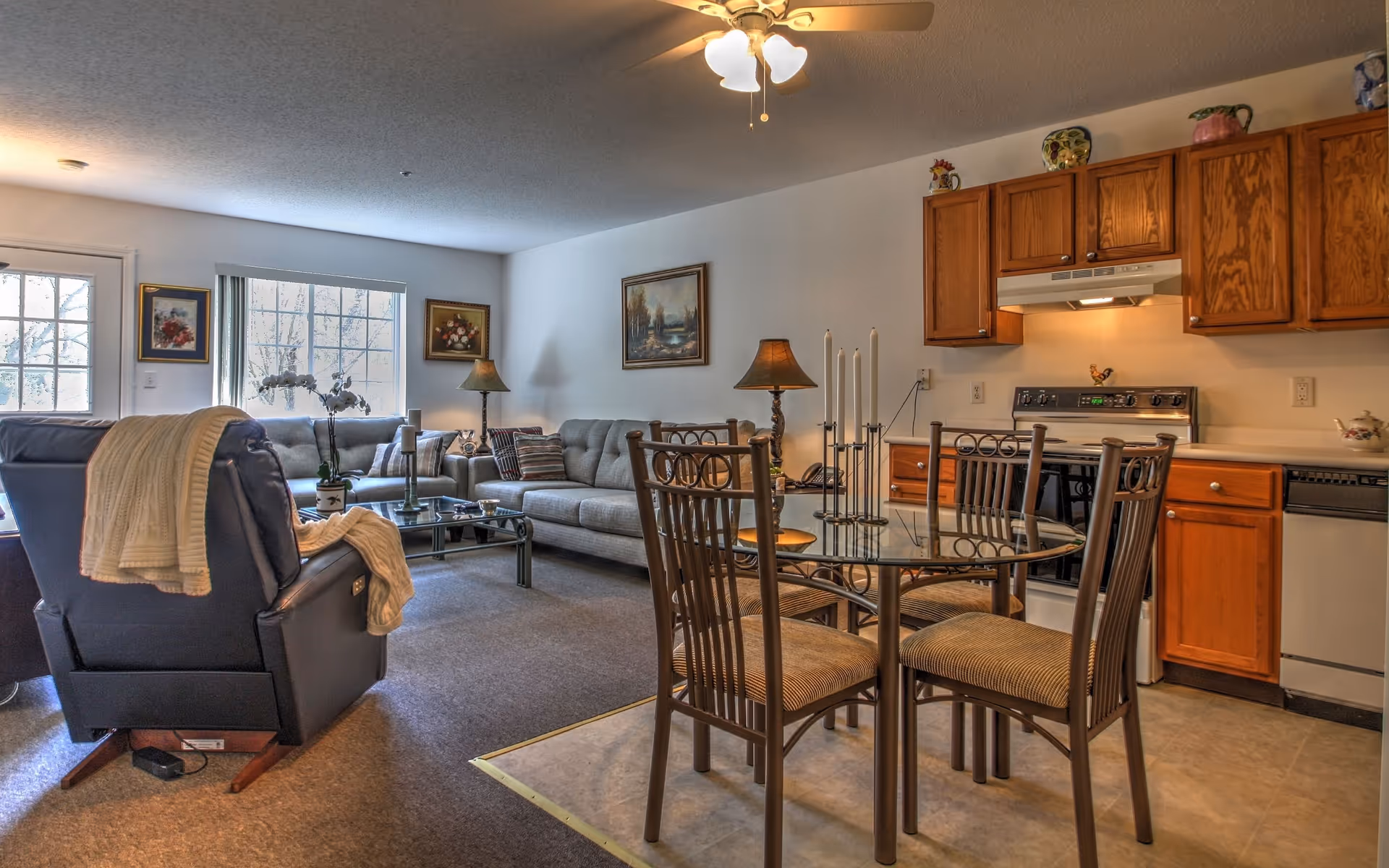 A cozy living and dining area in a senior living facility named Spruce Manor. The space features a glass dining table with four chairs, a kitchen area with wooden cabinets, a stove, and a dishwasher. The living area has a gray sofa, a black recliner with a cream-colored blanket draped over it, a glass coffee table with a potted orchid, two table lamps, and framed artwork on the walls. A ceiling fan with lights is mounted above.