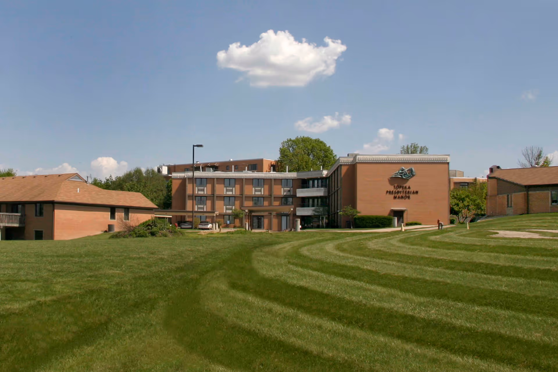 Exterior view of Topeka Presbyterian Manor, a multi-story brick building with a well-maintained lawn in the foreground and a partly cloudy sky above.
