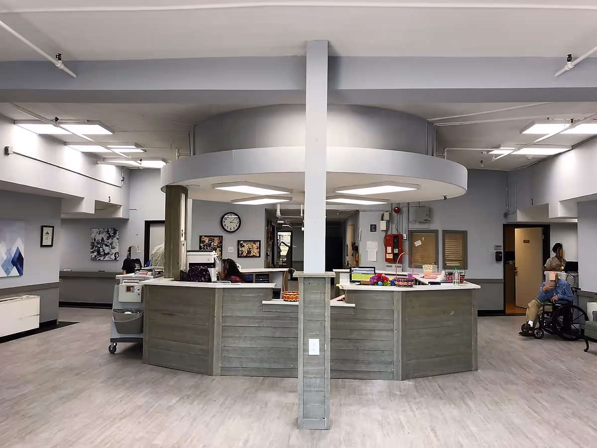 Interior view of a senior living facility reception or nurse station area with a circular desk in the center. Two staff members are seated behind the desk working, and a person in a wheelchair is visible on the right side. The room has light gray walls, ceiling lights, and some artwork on the walls.