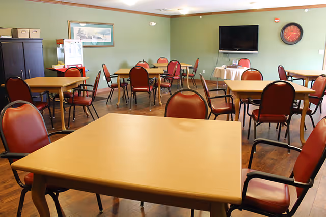 A dining room with multiple square wooden tables and red cushioned chairs arranged around them. The room has green walls, a wooden floor, a wall-mounted flat screen TV, a clock, and a framed picture on the wall. There is also a cabinet and a small refrigerator in the corner.