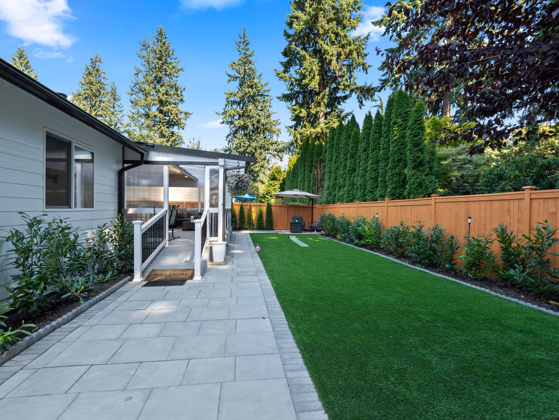 Outdoor backyard with a covered patio, paved path, green lawn, and privacy fence.