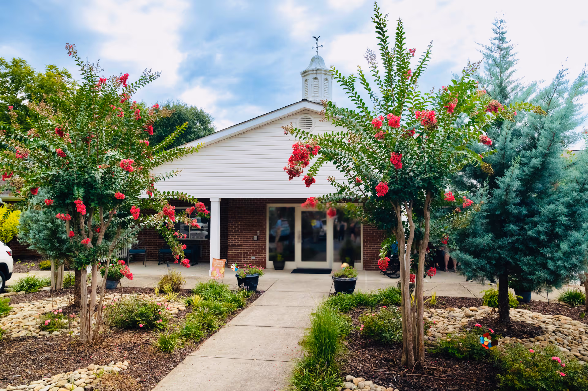 Front exterior view of a single-story building with a white gabled roof and brick walls, surrounded by landscaped garden beds with flowering trees and shrubs. A concrete pathway leads to the entrance with glass double doors.