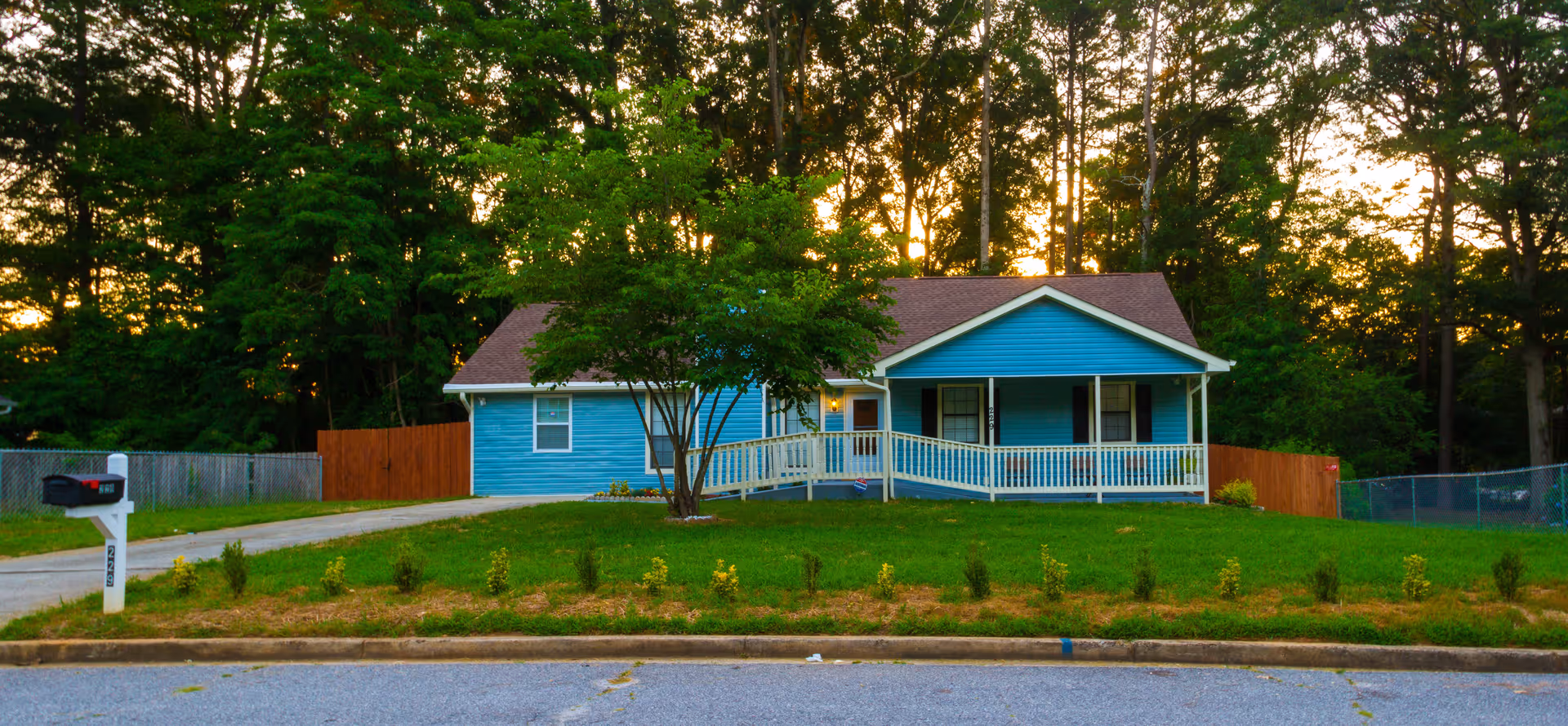 A single-story blue house with a brown roof, white porch railing, and a wheelchair ramp leading to the front door. The house is surrounded by green grass, small shrubs, and trees in the background during sunset. A white mailbox is visible near the street.