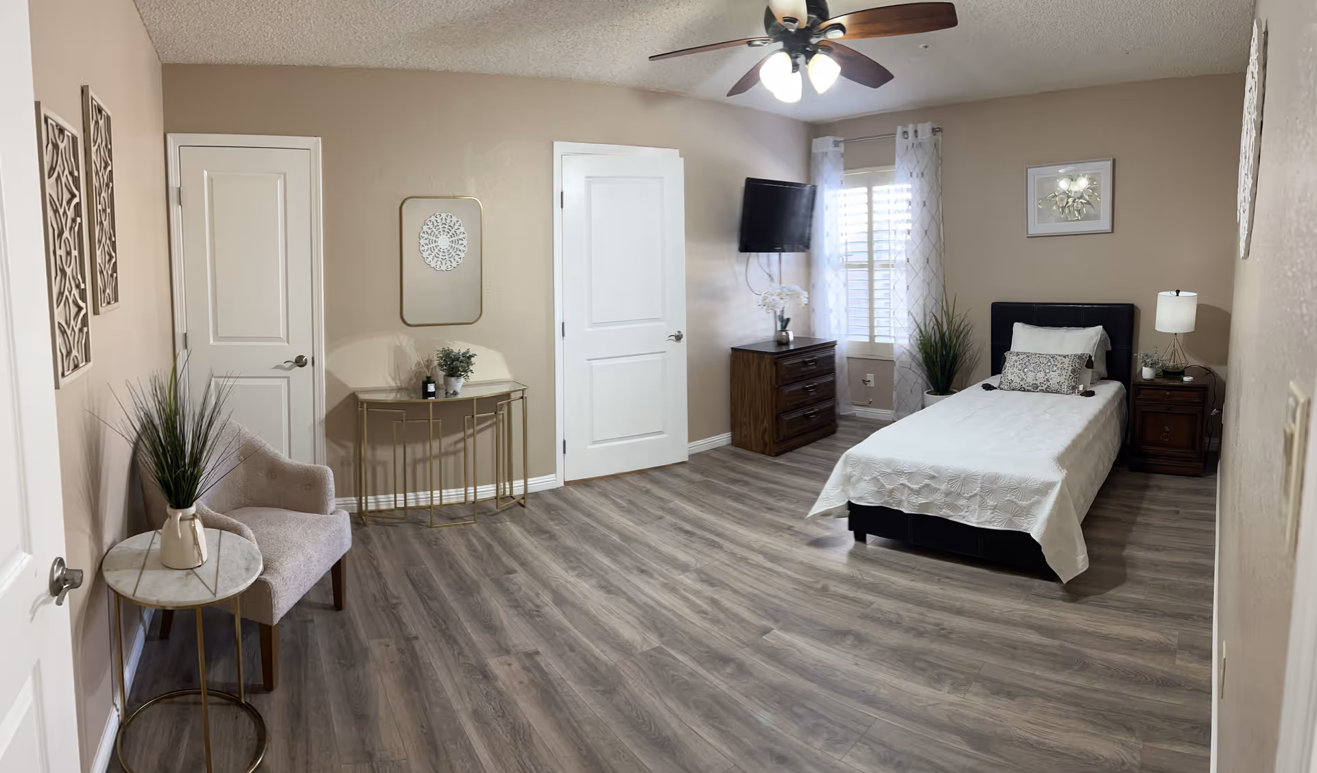 A spacious, well-lit senior living bedroom with bed, dresser, TV, and seating area.