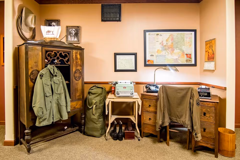 Vintage-themed interior nook with a wooden armoire, desk and chair, a typewriter, military jackets, map and framed pictures on the wall.