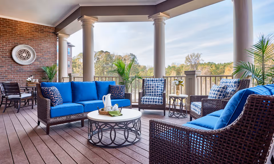 A spacious outdoor patio area with wicker furniture featuring blue cushions, including two sofas and two armchairs arranged around a round coffee table. The patio has wooden flooring, large columns, and a brick wall with decorative elements. There are potted plants and a scenic view of trees in the background under a partly cloudy sky.