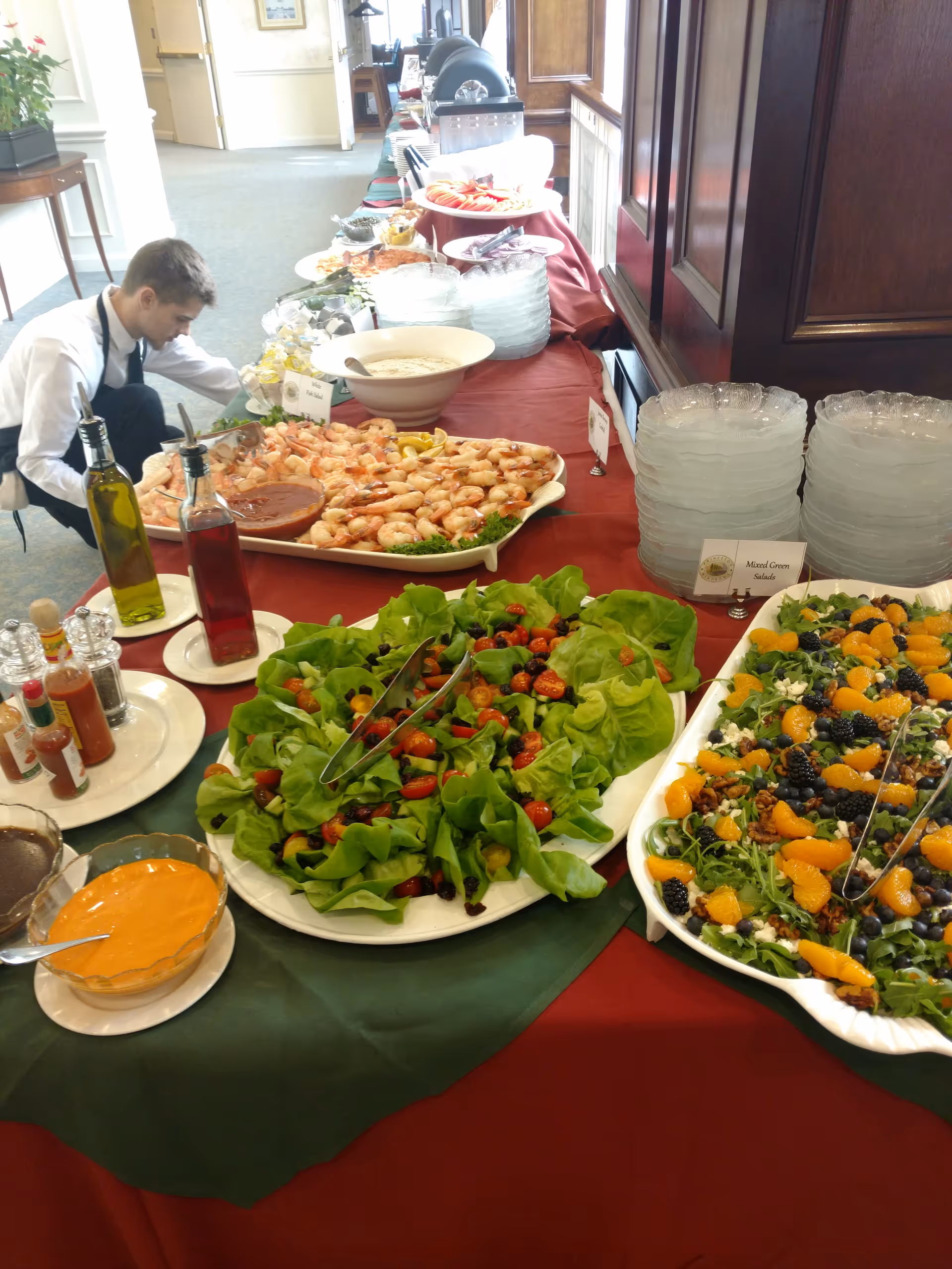 A staff member arranges a long buffet with salads, fruit, and sauces in a dining area.