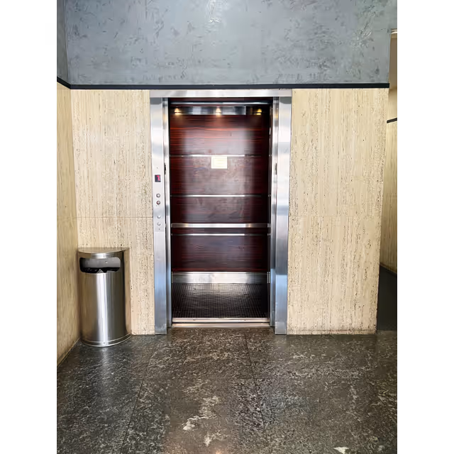 Elevator with metal frame in a beige-tiled hallway; trash can nearby.