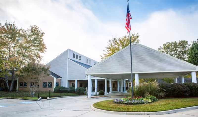 Exterior view of The Cottages at the Glen facility showing the entrance with a covered drop-off area supported by white columns, an American flag on a flagpole in front, surrounded by landscaped greenery and trees under a partly cloudy sky.