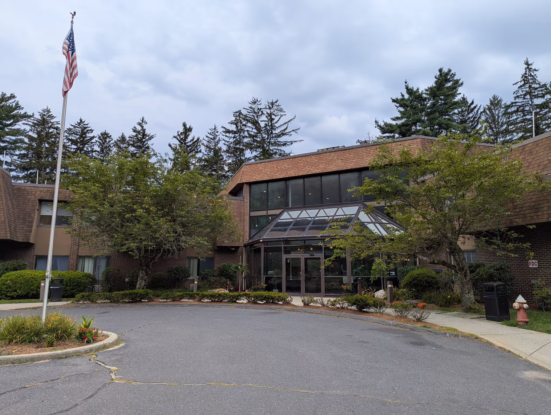 Front view of a brick senior living facility entrance with glass doors, trees, and a flag.