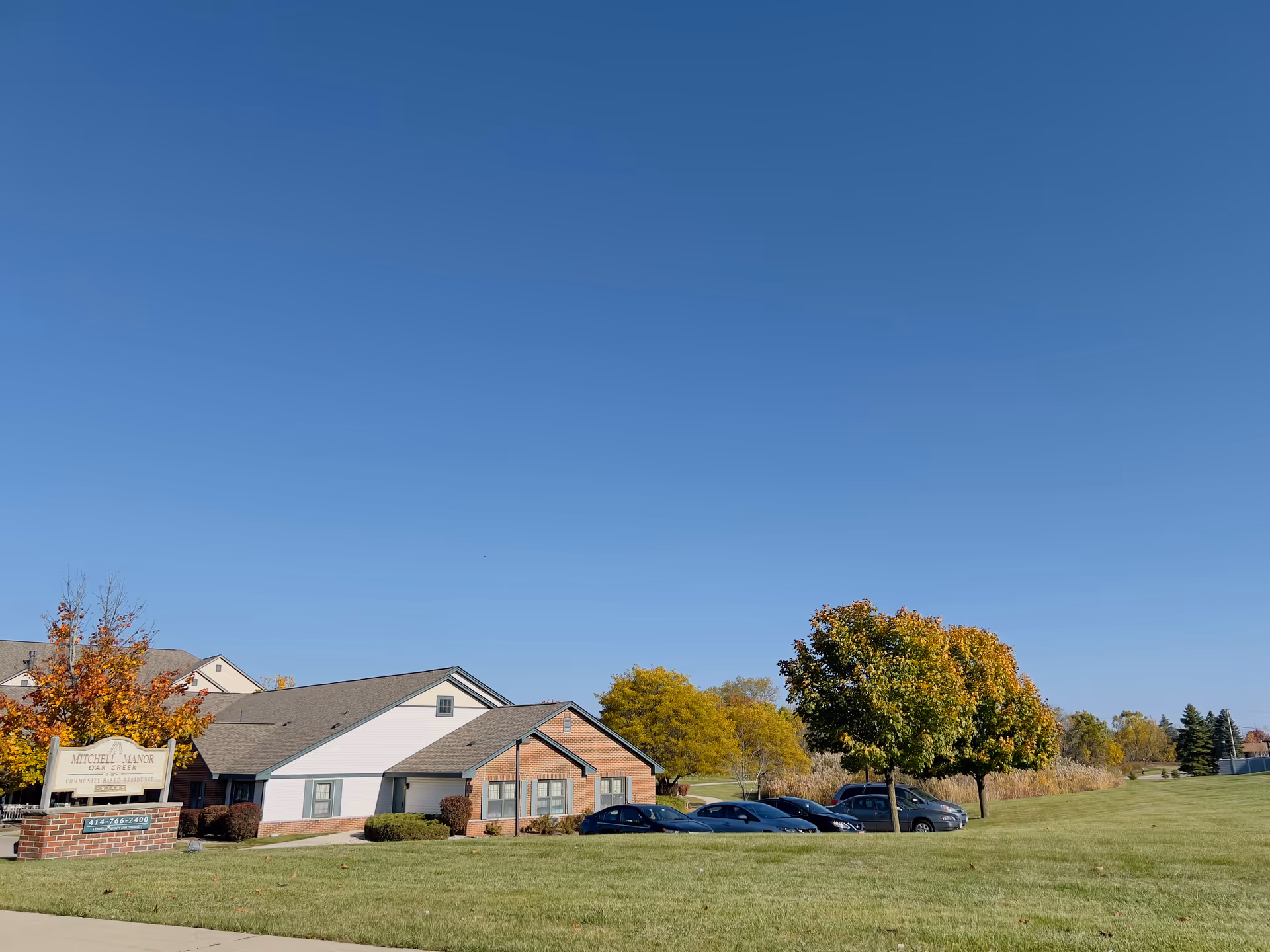 Exterior view of Mitchell Manor, a single-story brick and white siding building with a parking lot and several cars. There are trees with autumn foliage and a clear blue sky above.