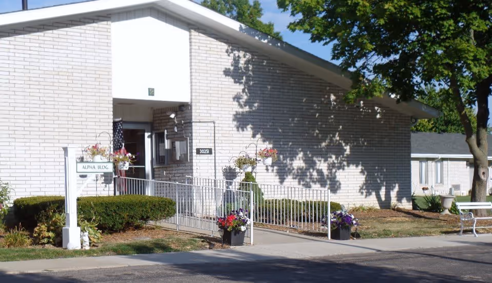 Entrance of The Baptist Manor Alpha Building showing a white brick façade, wheelchair ramp, sign and potted flowers.