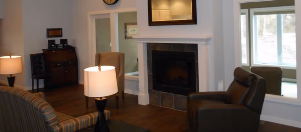 A cozy living room featuring a fireplace with a tiled surround and white mantel, a striped sofa, a dark leather armchair, two table lamps with white shades, and a wooden cabinet against the wall. The room has hardwood flooring and windows that allow natural light to enter.