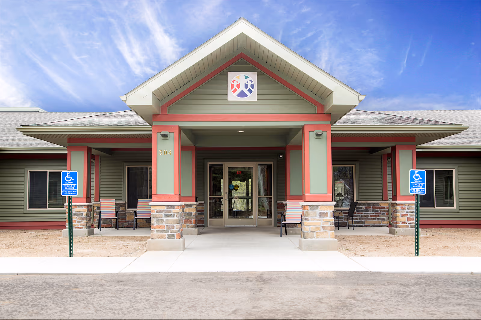 Front entrance of Carefree Living Aurora facility featuring a covered walkway with stone and green siding, two handicap parking signs, and several chairs on the porch under a blue sky with wispy clouds.