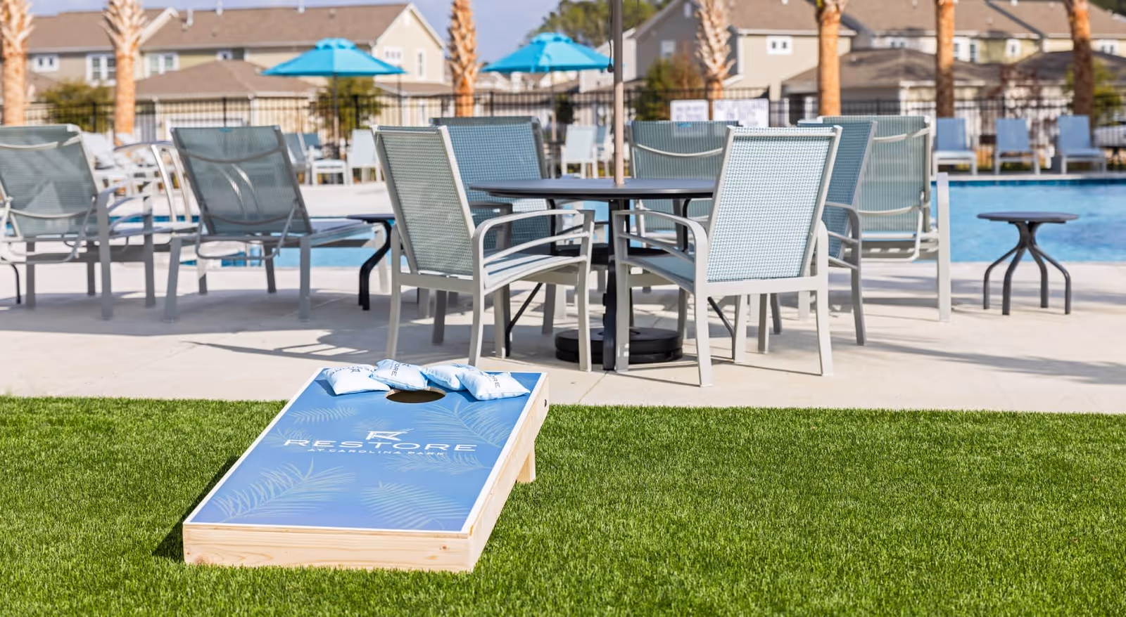 Outdoor poolside seating area with a cornhole board on artificial turf in front of tables, chairs, and a swimming pool.