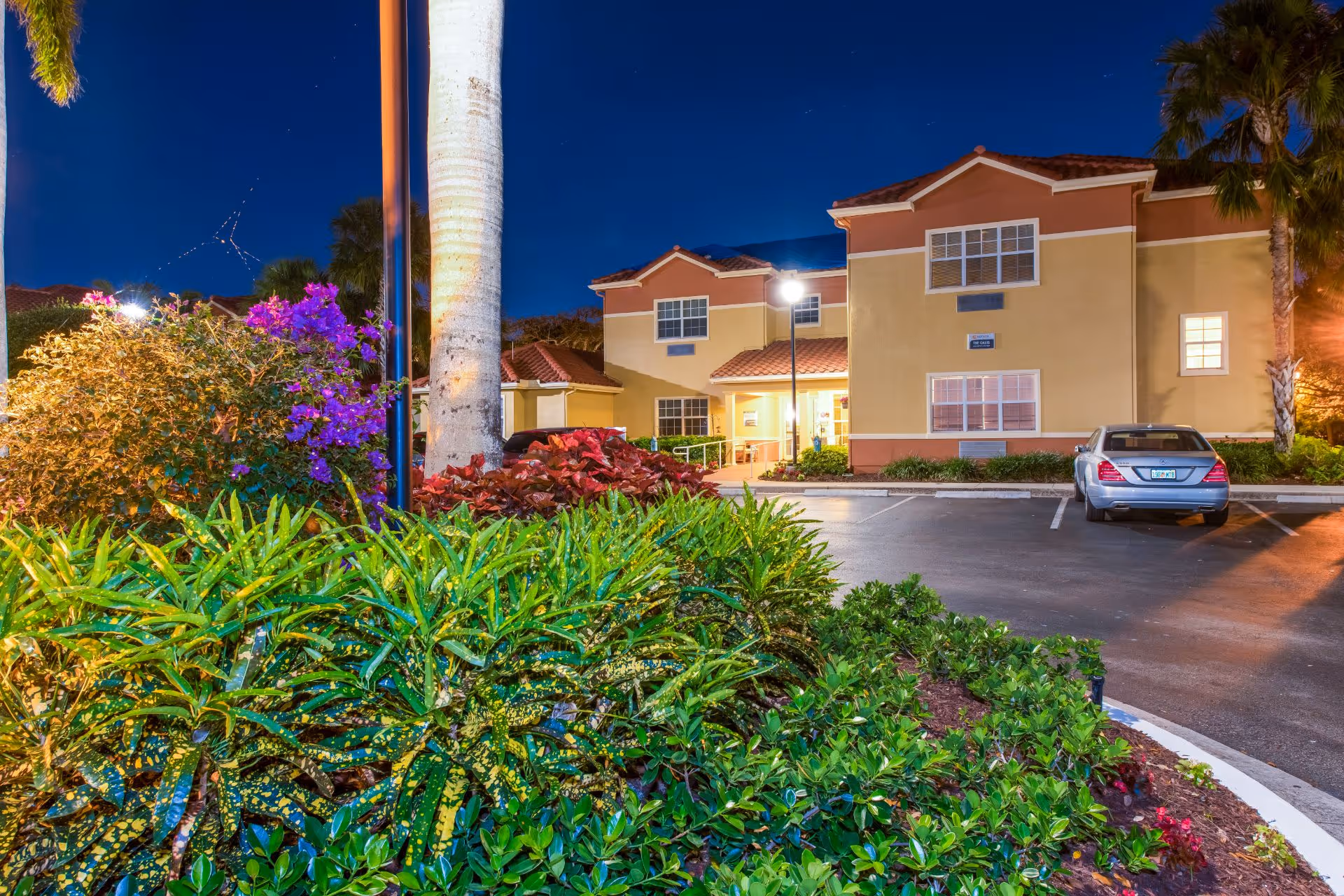 Front exterior of Sonata Delray Beach at night with landscaped greenery, a parking lot and a parked car.