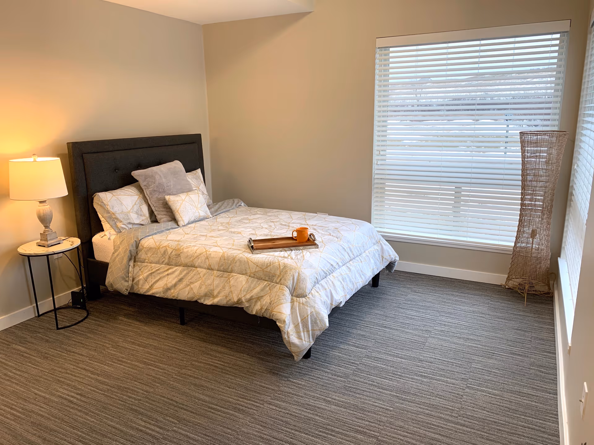 A well-furnished bedroom featuring a bed, bedside table, lamp, and a window with blinds.