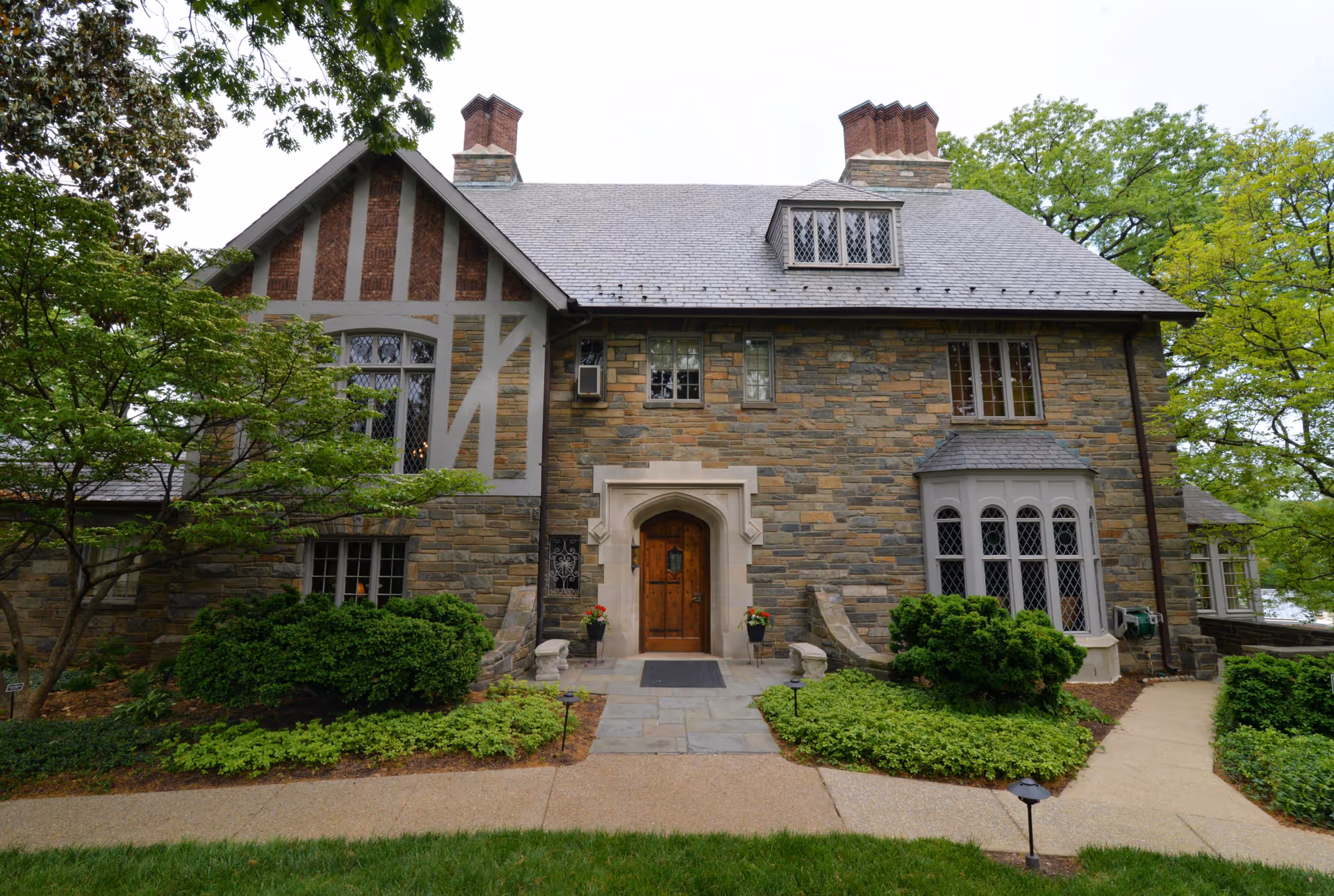 Stone Tudor-style building facade with a central wooden entrance, leaded windows, chimneys, and landscaped walkway.
