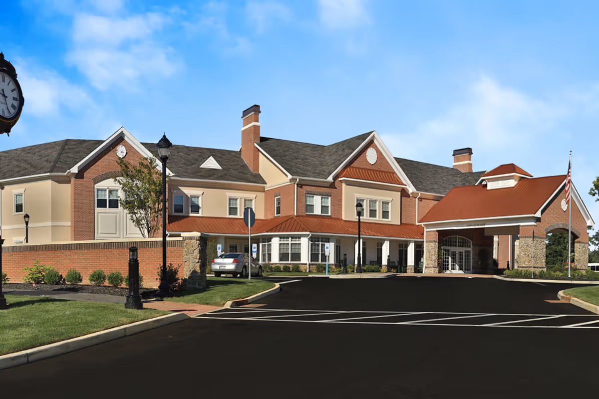 Front exterior view of Brandywine Voorhees by Monarch, a two-story building with a combination of brick and beige siding, a red roof over the entrance, several windows, a clock on a pole to the left, and a flagpole with an American flag on the right. There is a driveway and parking area in front of the building with some landscaping.