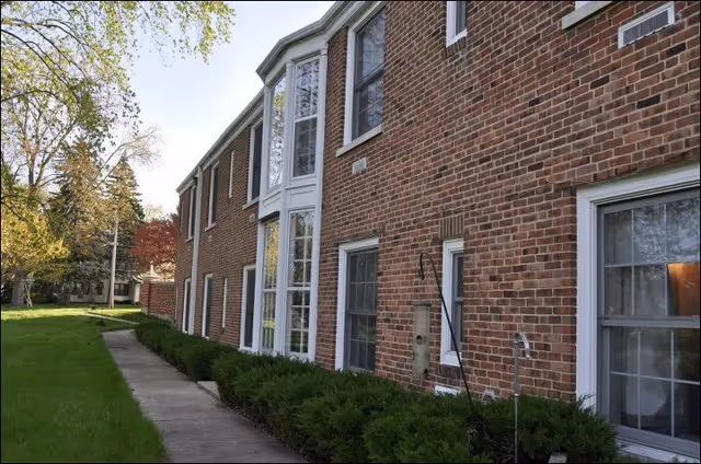 Brick senior living facility exterior with large windows, a sidewalk path, and neatly trimmed shrubs.