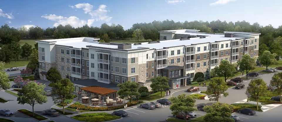 A large, modern four-story senior living facility surrounded by trees and greenery. The building features multiple balconies, a stone and light-colored exterior, and a covered entrance. There is a parking lot with several cars and a patio area with tables and umbrellas in front of the building.