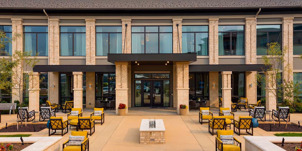 Outdoor seating area and main entrance of a two-story brick senior living building with yellow chairs and a central walkway.