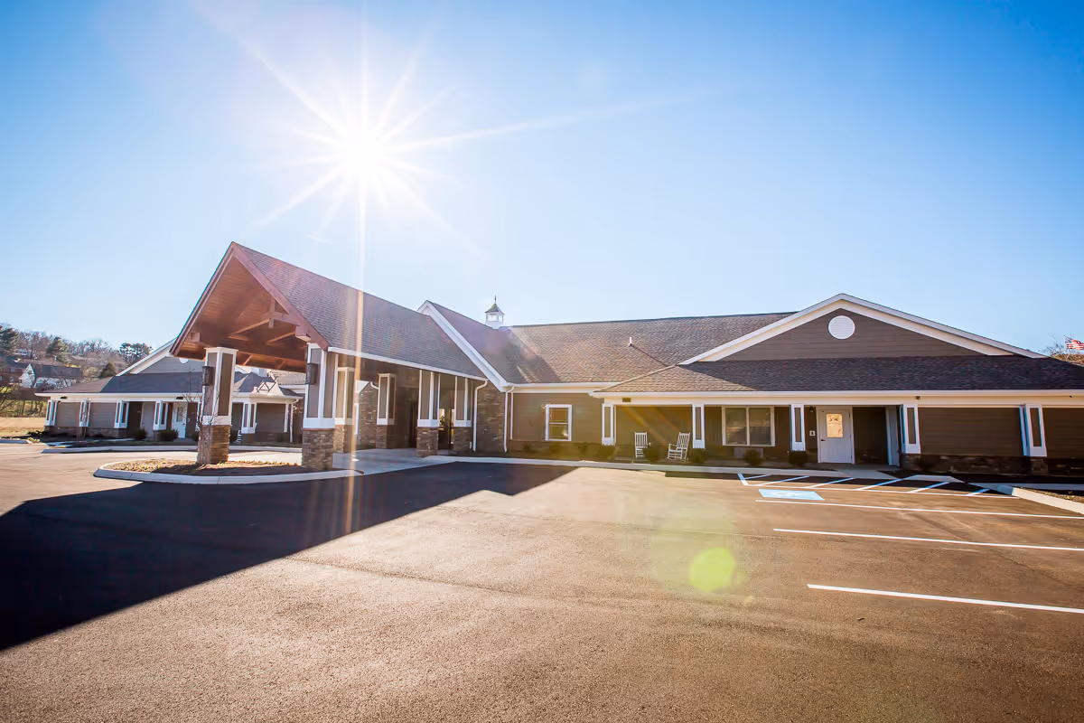Exterior view of a single-story senior living facility building with a covered entrance, stone pillars, and a large parking lot under a bright sunny sky.