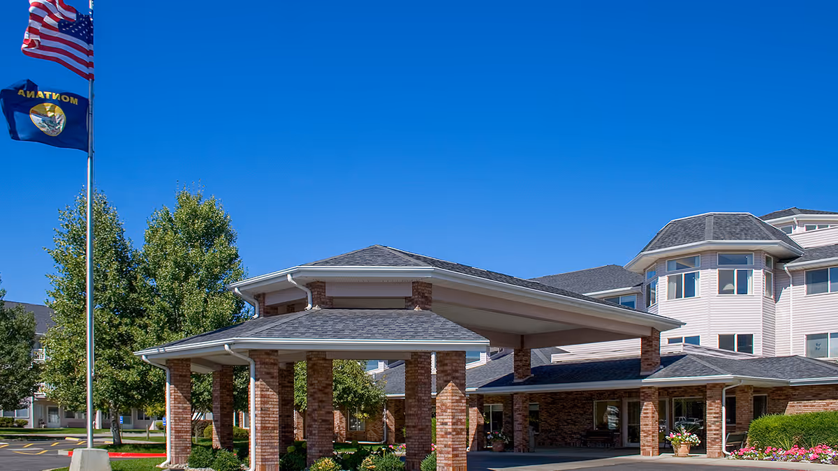Exterior view of Holiday Aspen View facility showing a covered entrance with brick pillars, a multi-story building with many windows, and two flagpoles displaying the American flag and the Montana state flag under a clear blue sky.