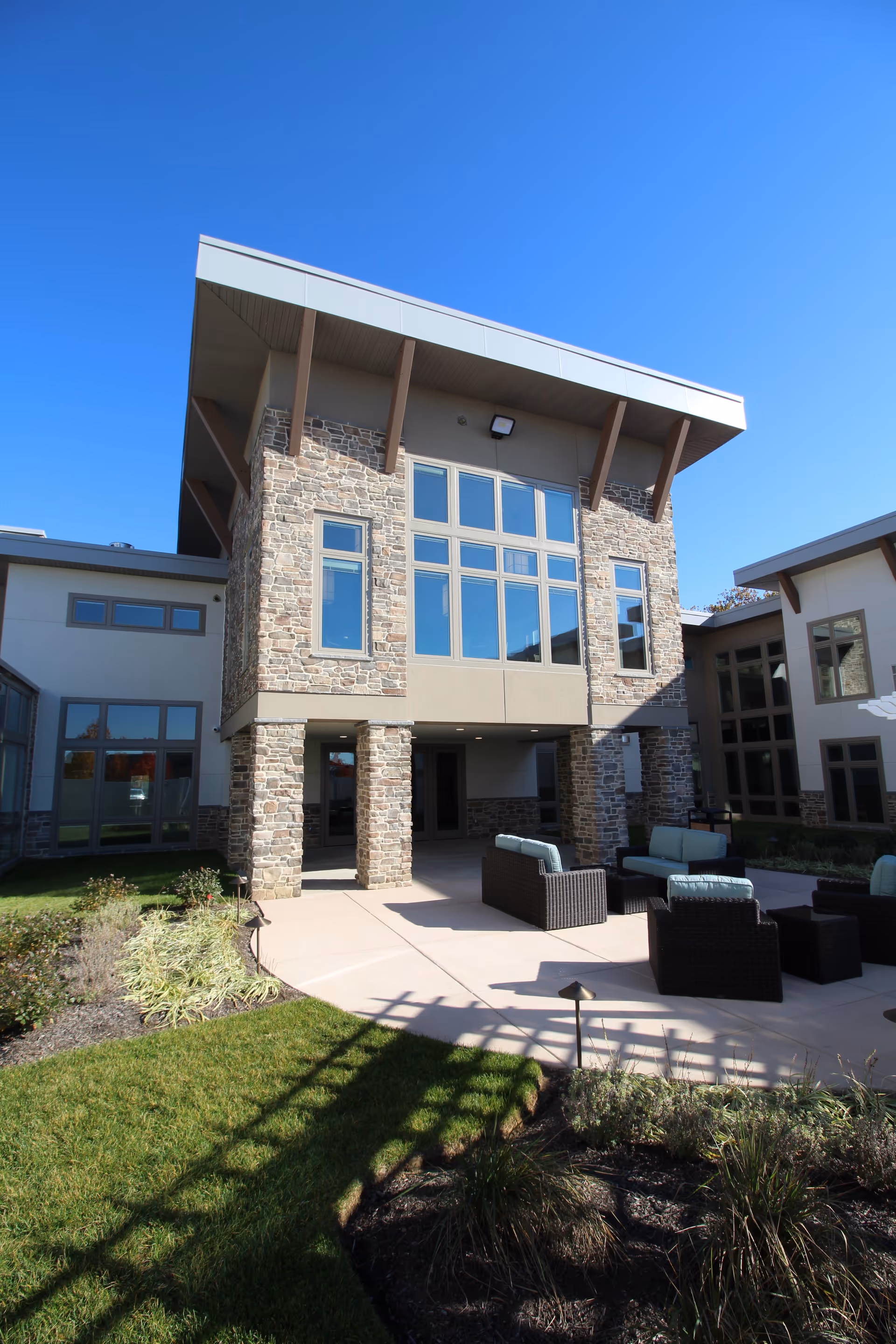 Stone-front senior living building with large windows and an outdoor patio furnished with wicker seating under a clear blue sky.