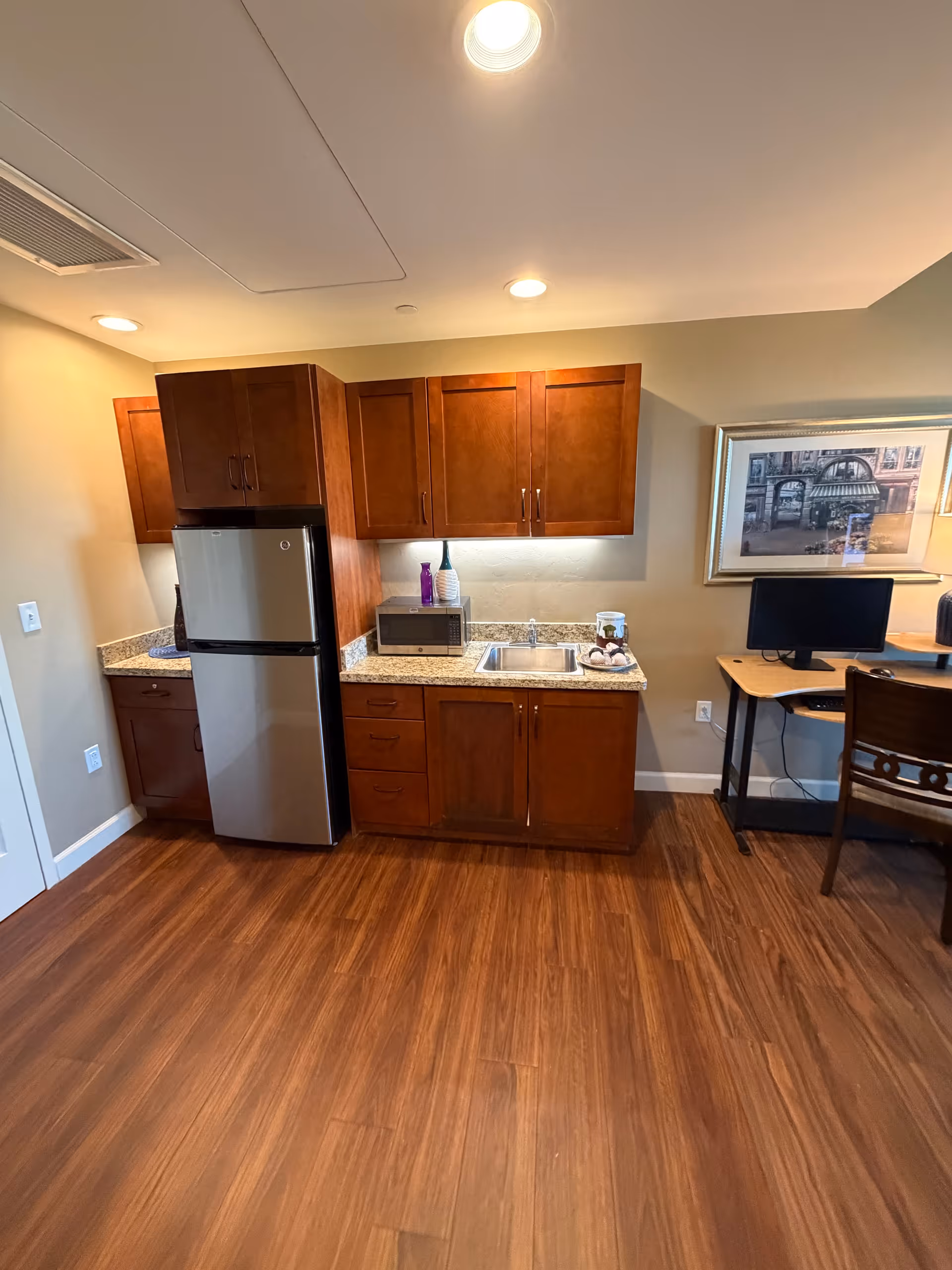 Wooden cabinets and a stainless steel fridge line a kitchenette with sink, microwave, and computer desk.