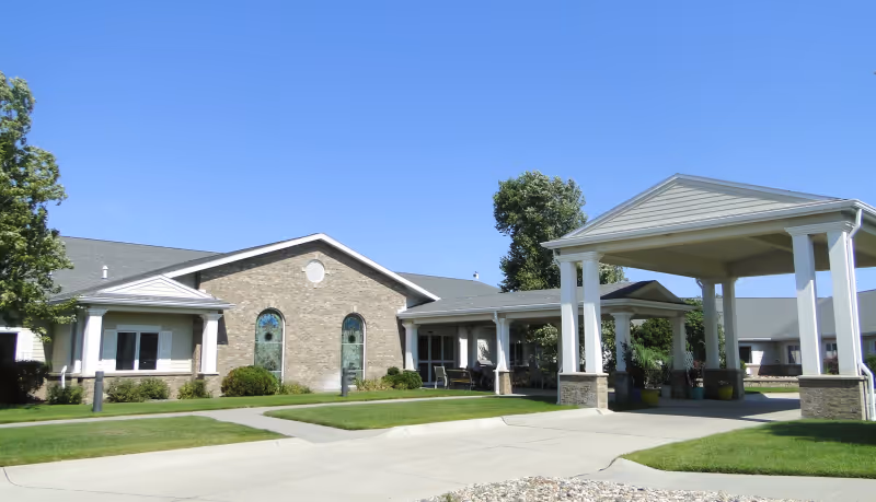 Front exterior of a single-story senior living facility with a covered entrance porte-cochère, brick facade, and landscaped lawn under a clear blue sky.