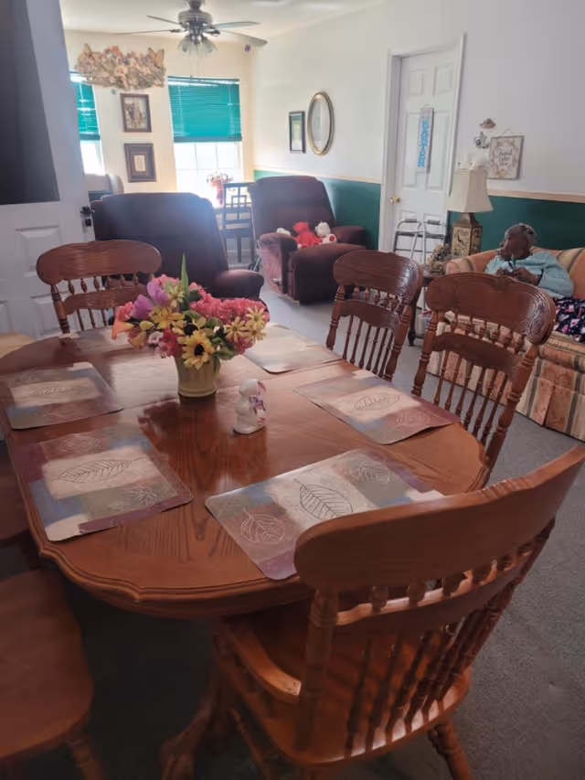 A wooden dining table with chairs in a senior living facility; lounge area visible in background.