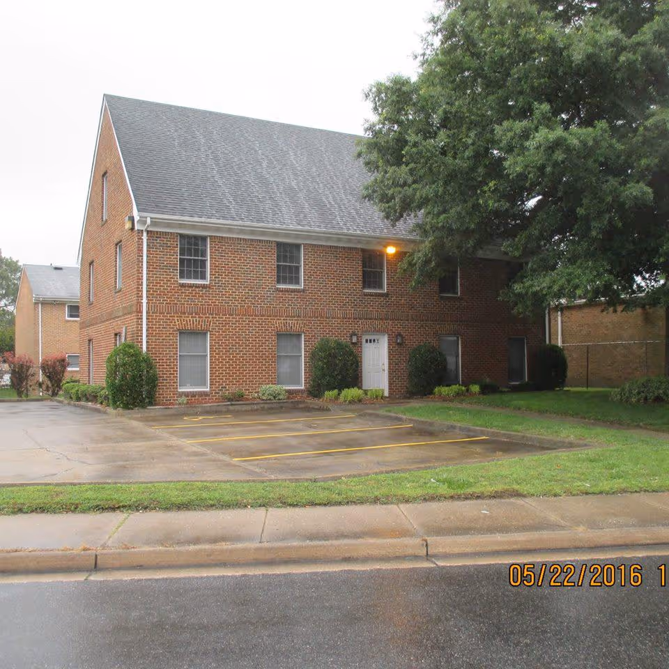 Two-story brick building with a parking lot, shrubs, and a large tree.